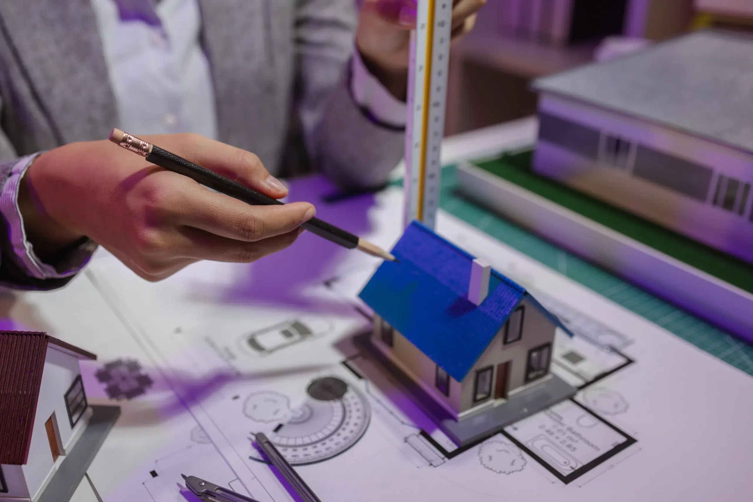 A person measures a small house model with a ruler on architectural plans, holding a pencil, symbolizing property appraisal and valuation in a condemnation case.