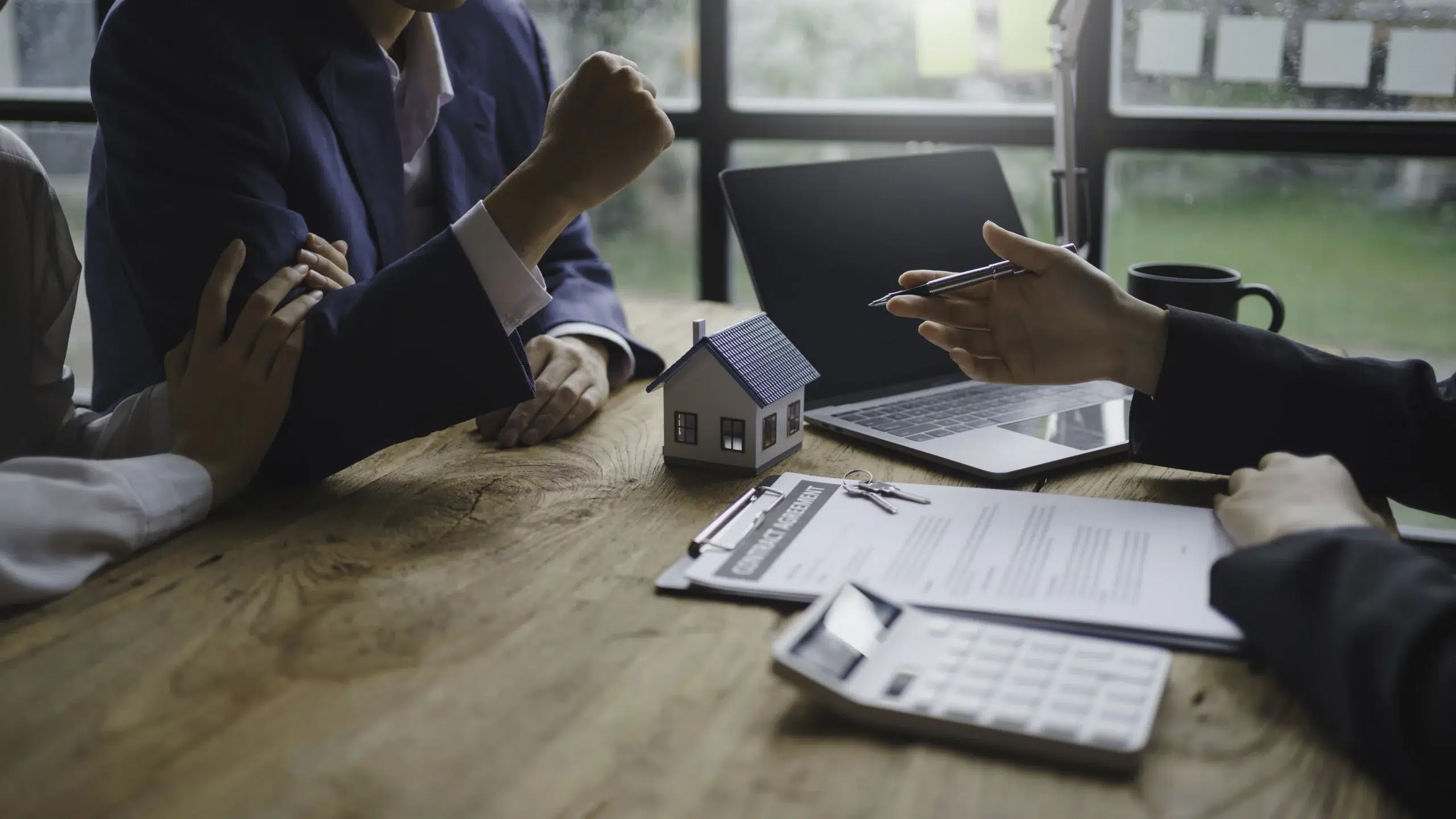 Two people discuss over a wooden table with a small house model, contract, and keys, symbolizing negotiations for government property acquisition