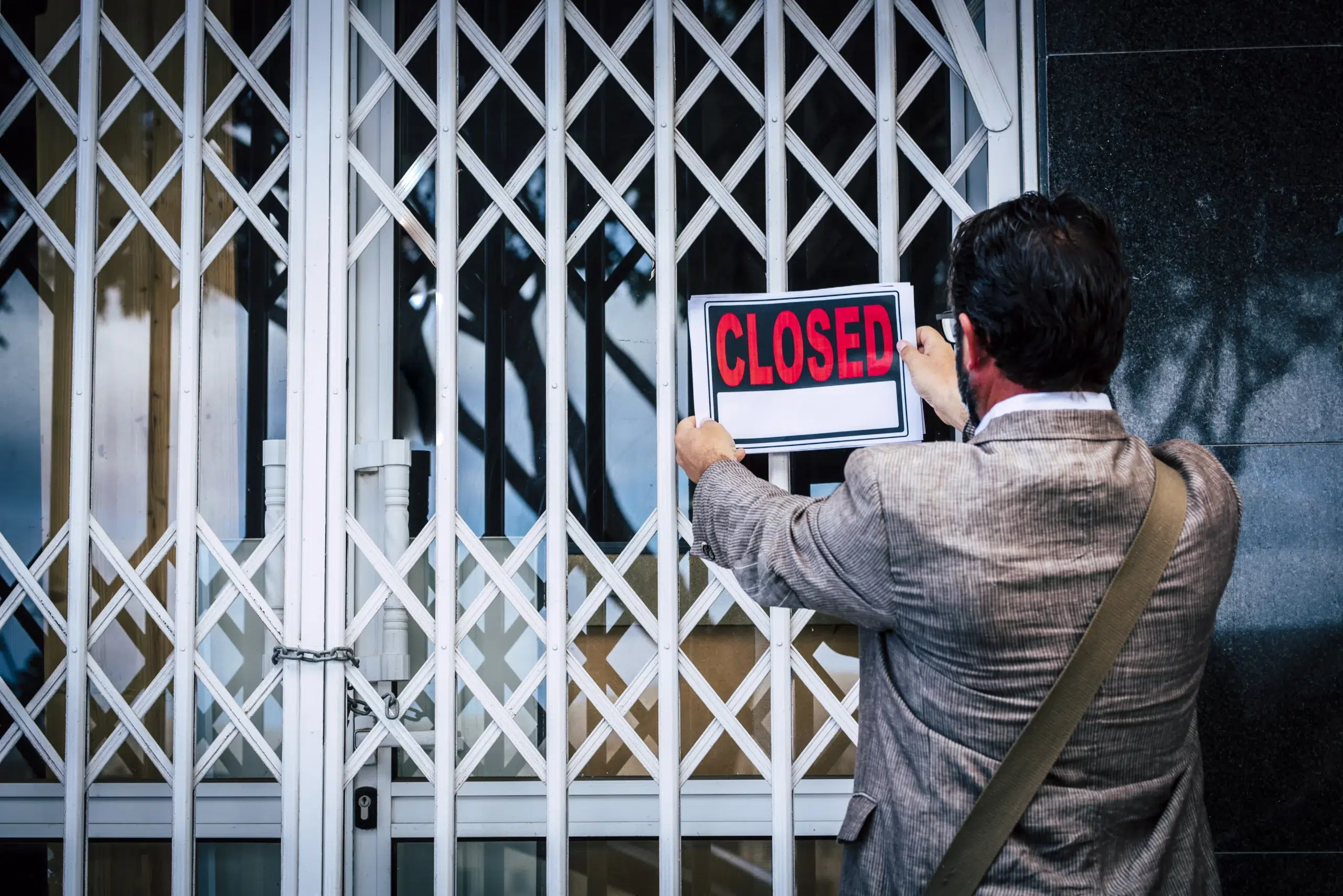 Man in suit hangs a closed sign on a locked gate with chain and padlock, symbolizing business shutdown or government seizure.